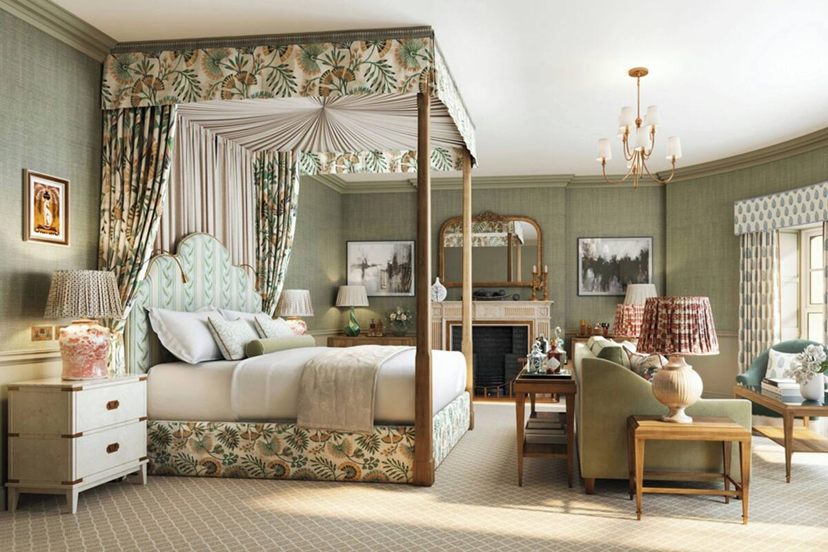 A four-poster canopy bed with floral upholstery, a sitting area with a green armchair and a wooden side table, and a fireplace with a mirror above it.