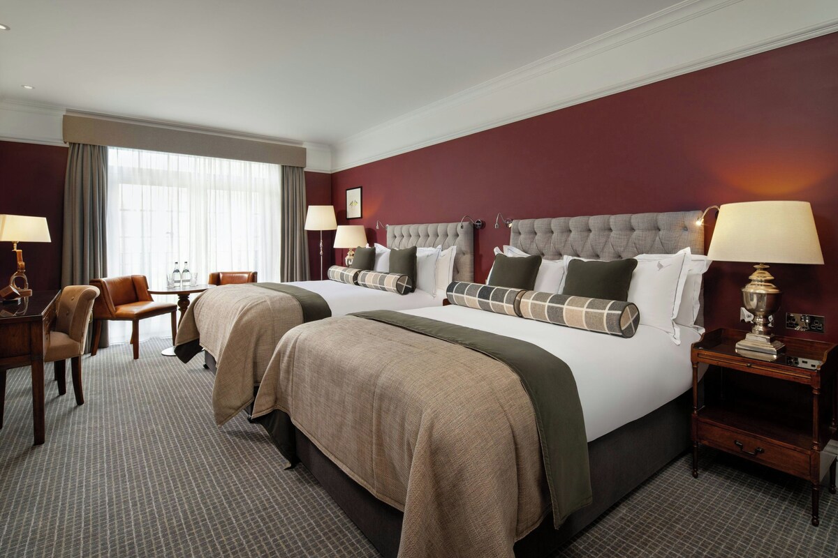 A hotel room with two beds, a desk, a chair, and a lamp.