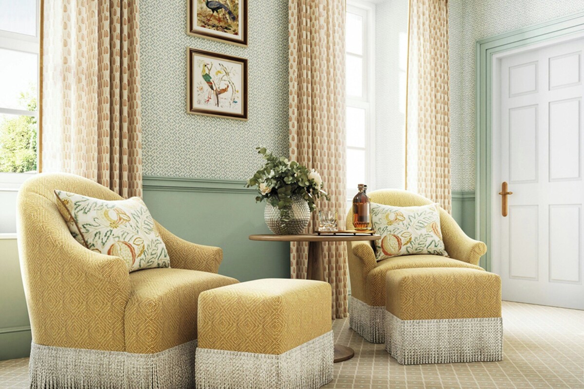 A room with two armchairs and ottomans, a small table with a vase and candle, and a door with a cross.