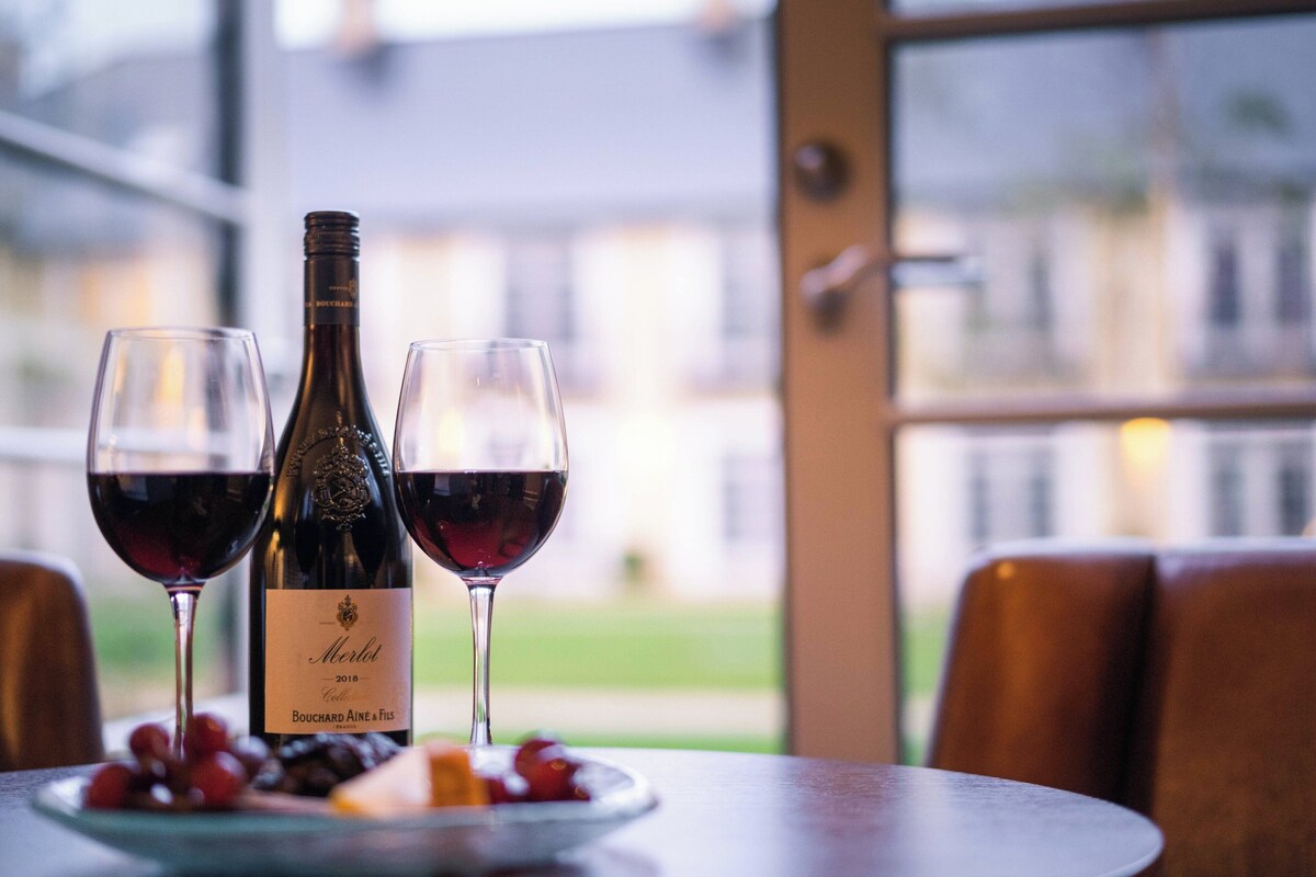 A table with a bottle of Merlot wine, two wine glasses, and a plate with grapes and cheese.
