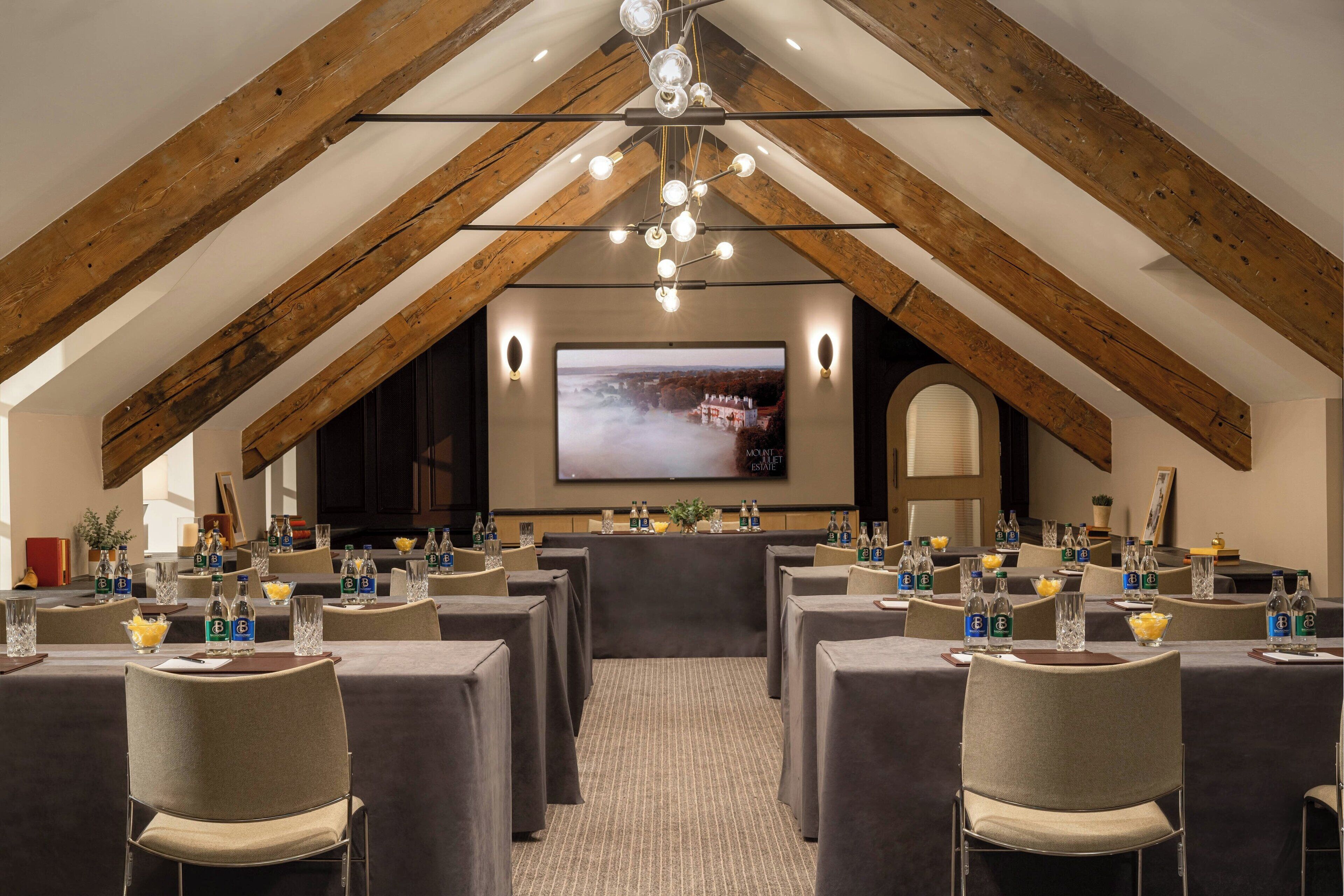 A conference room with a high ceiling and exposed wooden beams, equipped with tables, chairs, and a large screen for presentations.