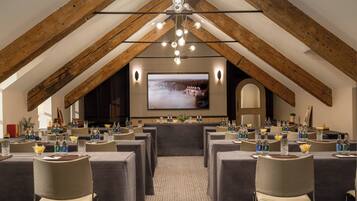 A conference room with a high ceiling and exposed wooden beams, equipped with tables, chairs, and a large screen for presentations.
