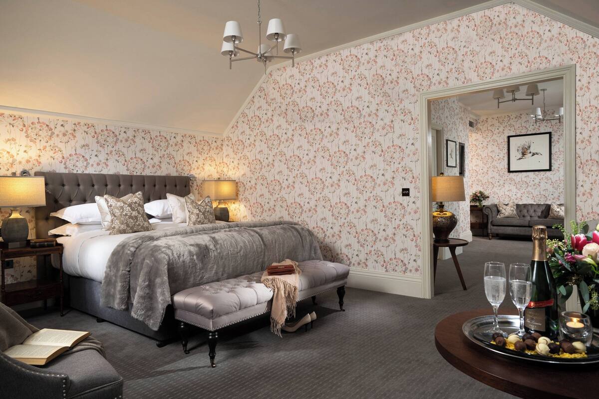 A well-appointed bedroom with a large bed, bedside tables, a reading chair, and a dining area with a tray of chocolates and a bottle of champagne.