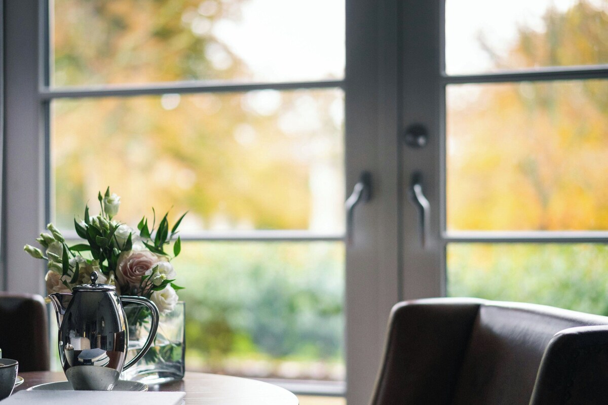 A table with a clear glass pitcher and a metal teapot, a vase with flowers, and a view of greenery through a window.