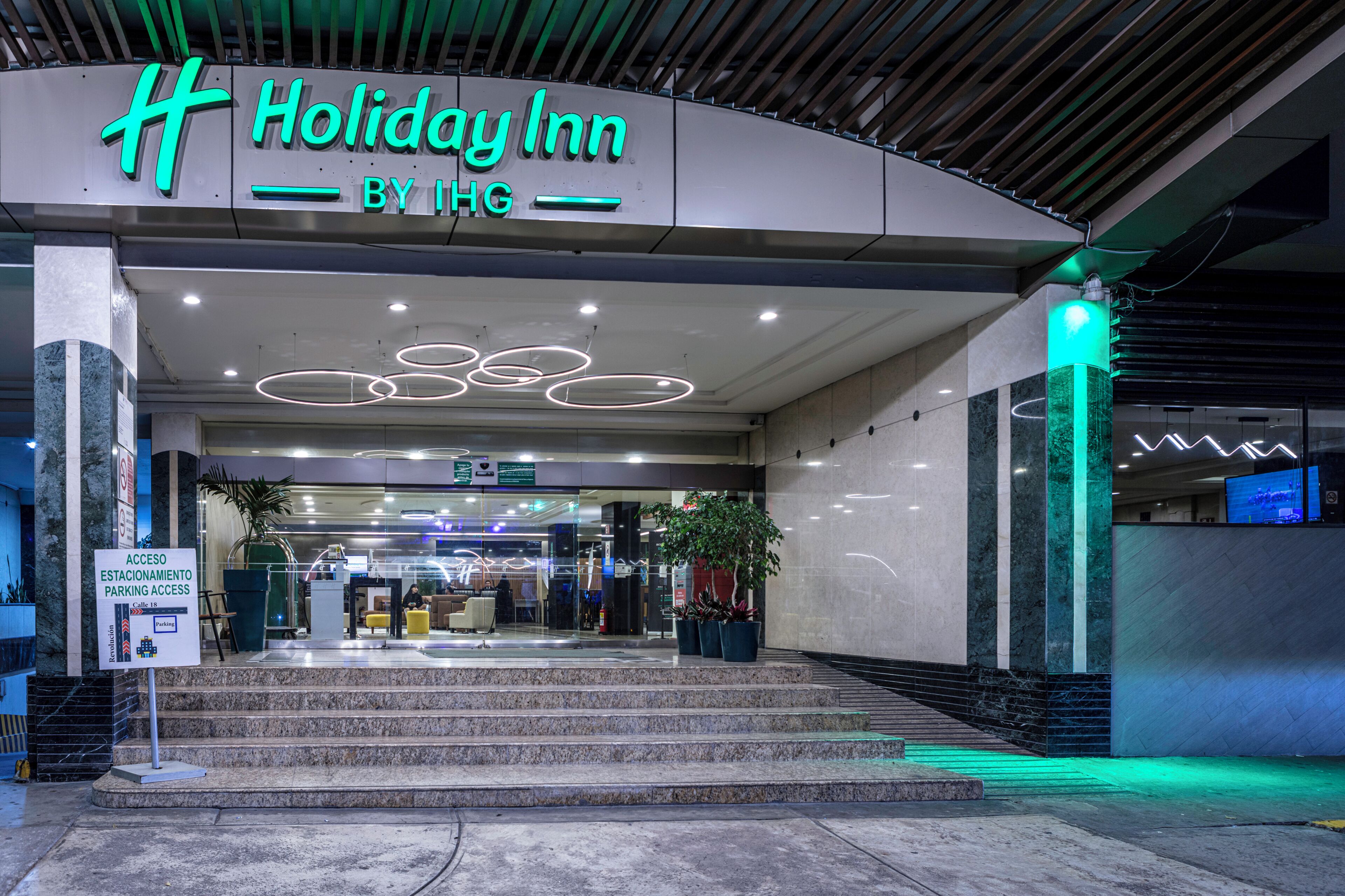 The exterior of Holiday Inn by IHG with a modern design, steps leading to the entrance, and a sign indicating parking access.