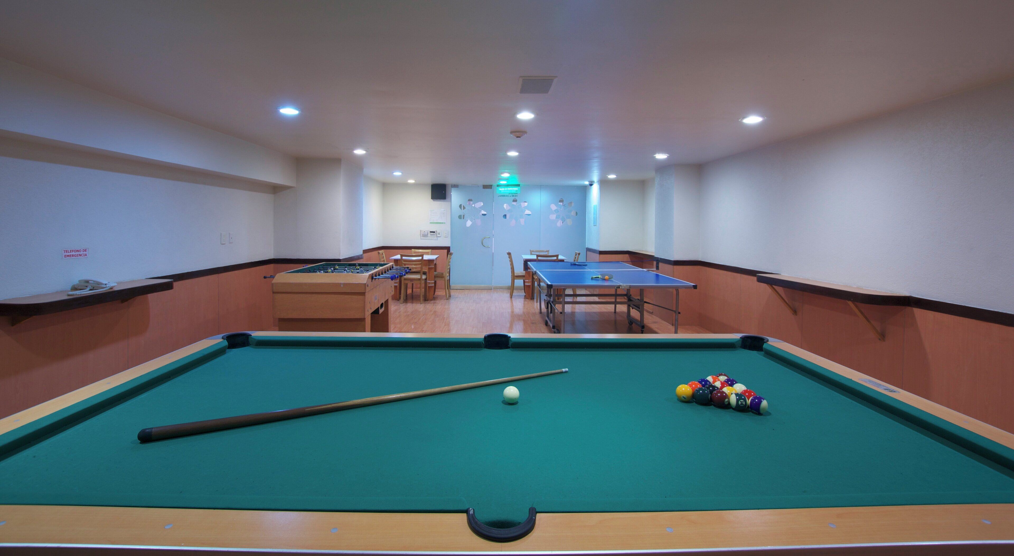 A pool table with a cue stick and balls, a foosball table, and a ping pong table in a room with wooden paneling and recessed lighting.