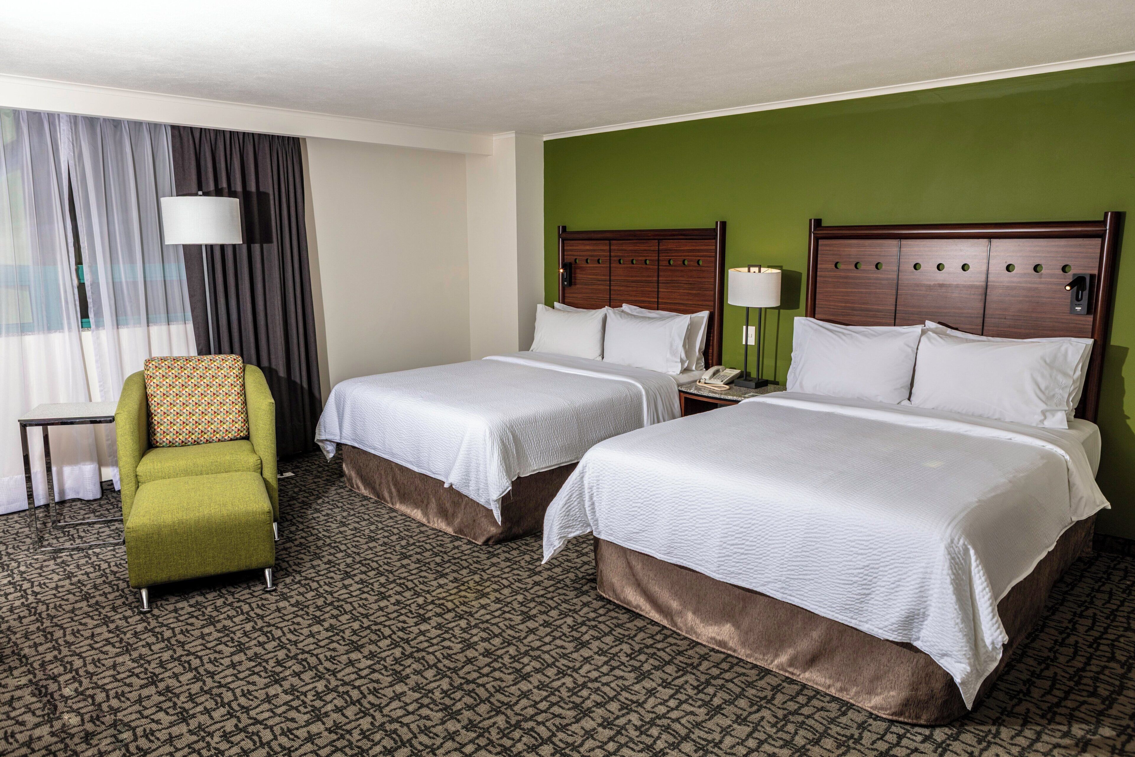 A hotel room with two beds, a green armchair, a desk, and a lamp.