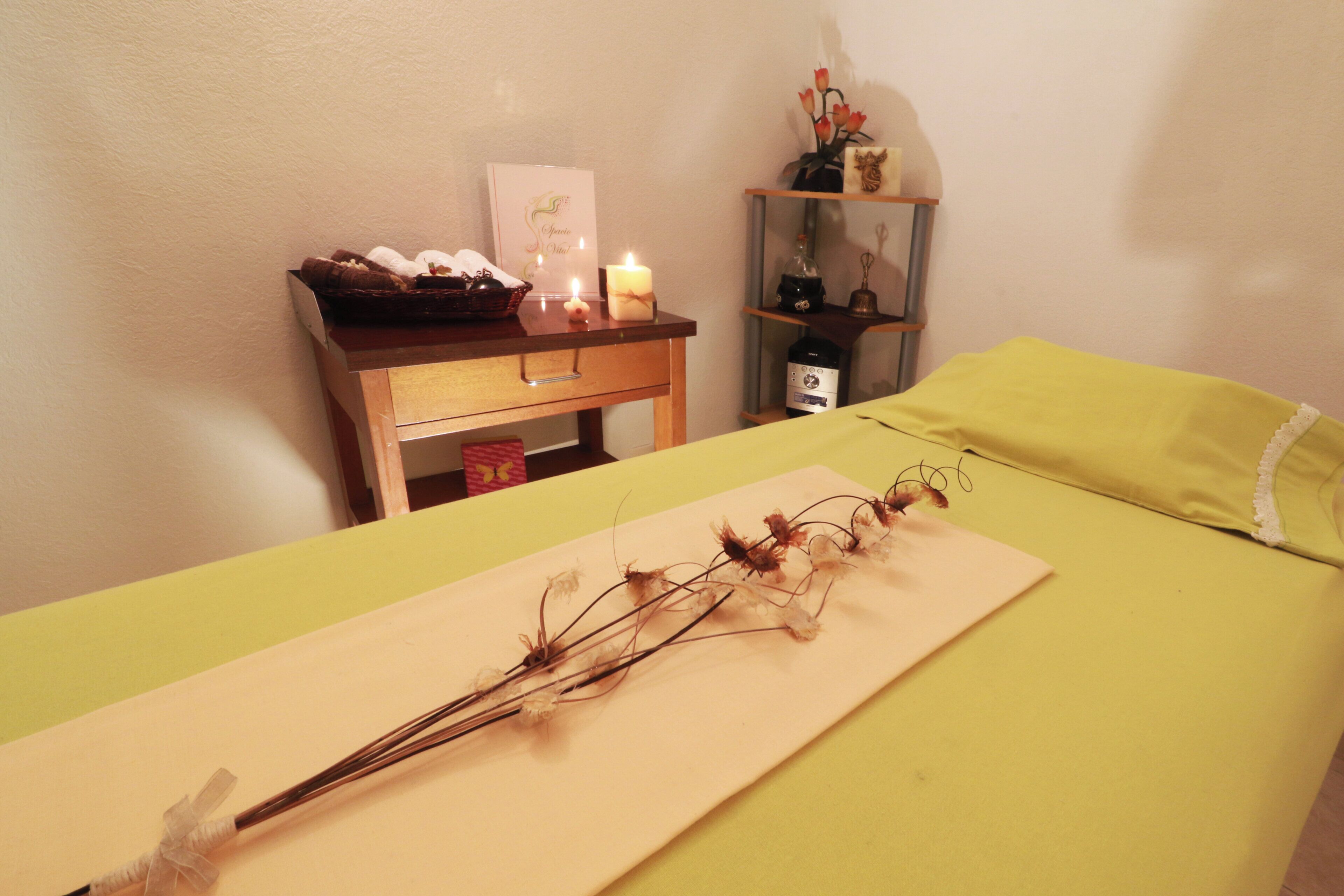 A room with a bed, a bedside table with a candle, a small shelf with decorative items, and a wall-mounted shelf with a bottle and a bell.