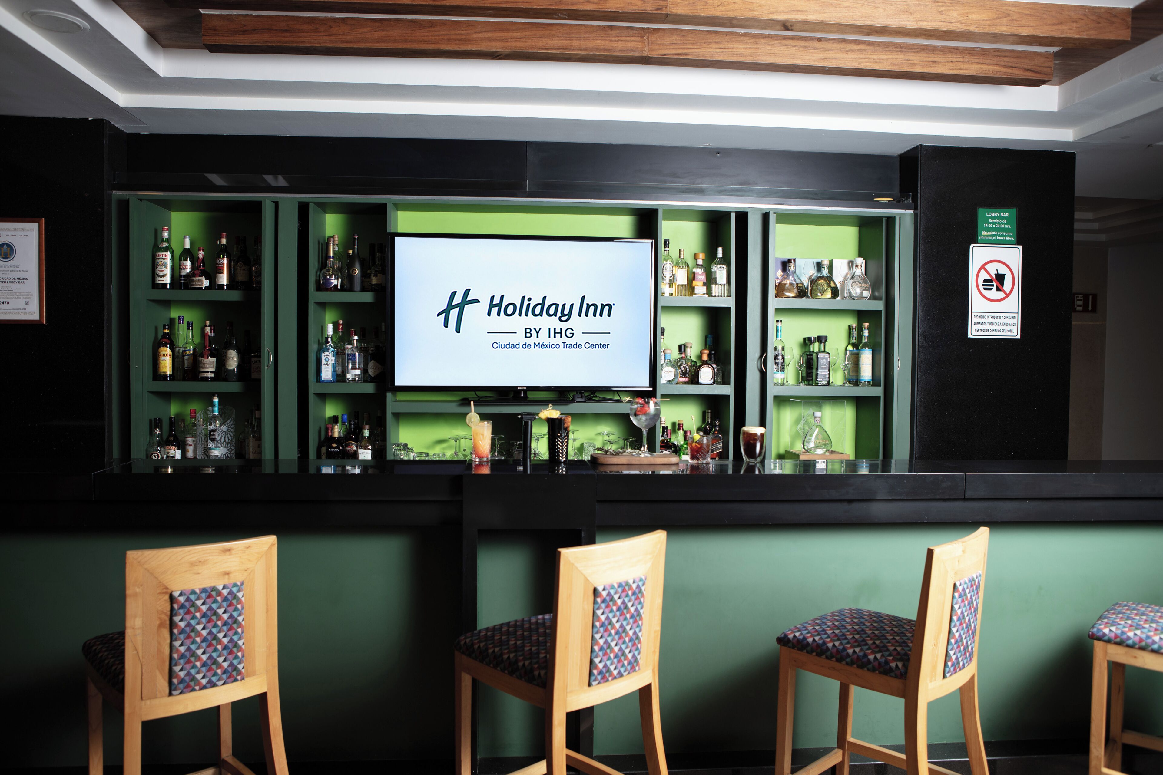 A bar area with green shelves, wooden chairs with patterned cushions, and a large screen displaying the Holiday Inn logo.