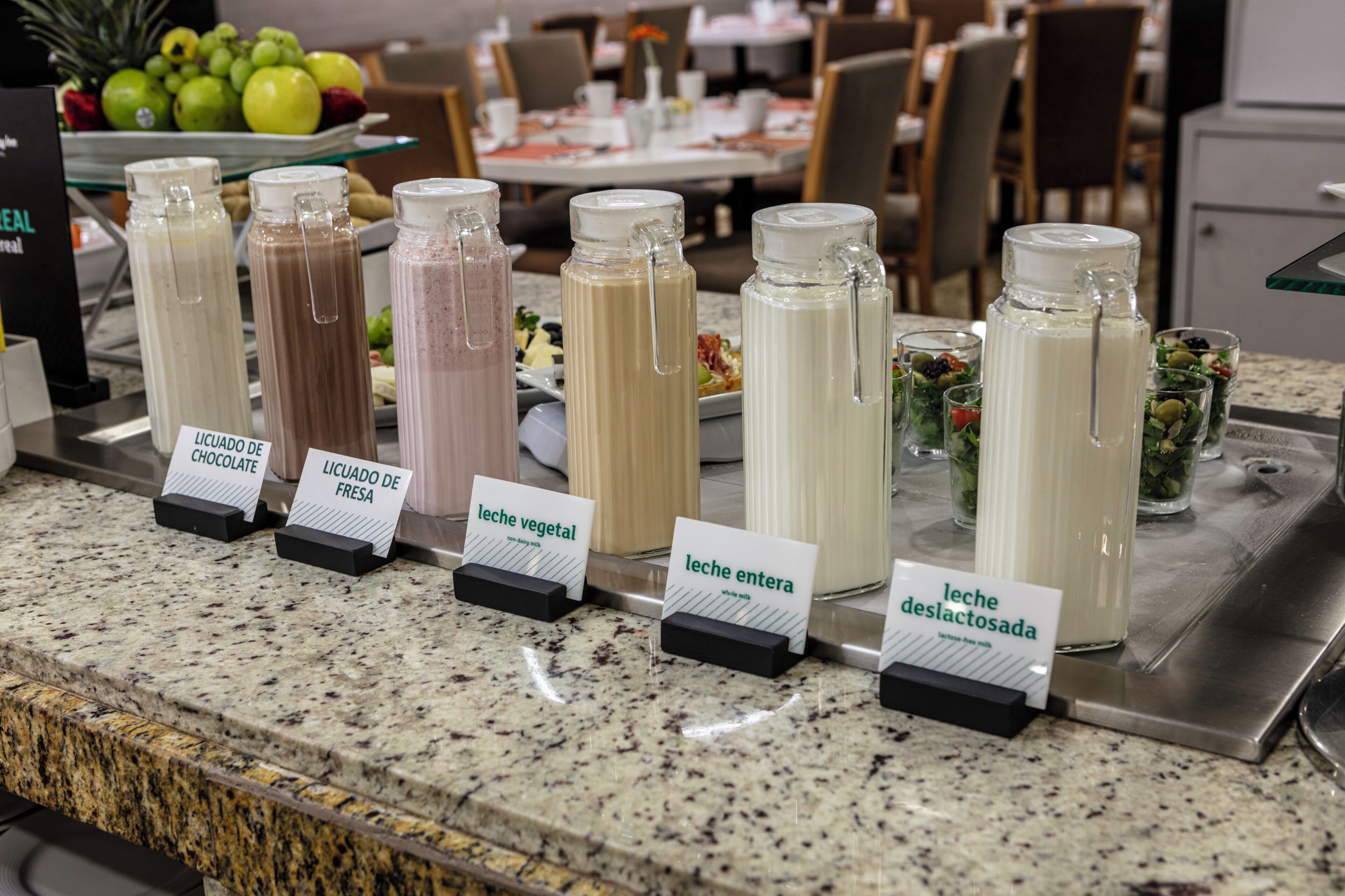 A beverage station with labeled drink options and a fruit bowl in the background.