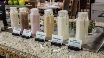 Una estación de bebidas con opciones claramente identificadas y un bol de frutas al fondo.
