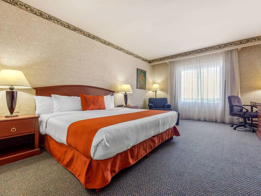 Een hotelkamer met een groot bed, twee nachtkastjes met lampen, een bureau met stoel en een groot raam met gordijnen.