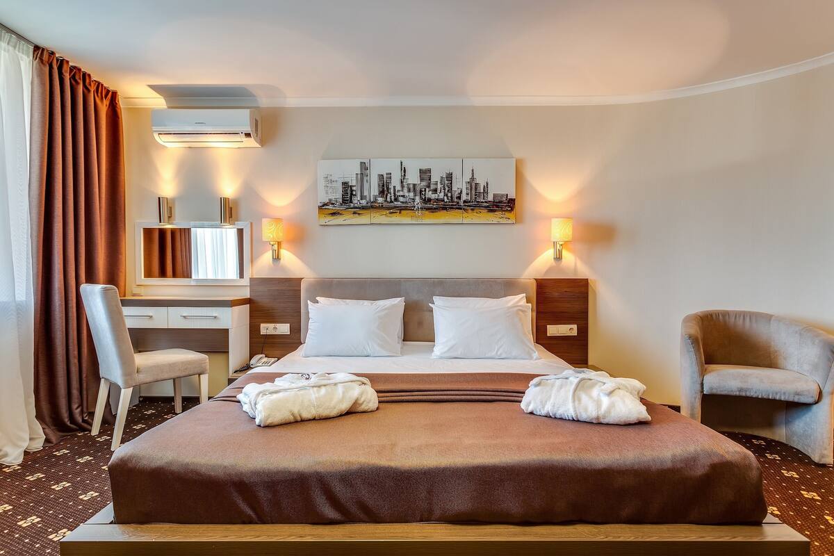 A hotel room with a bed, a desk, a chair, a cityscape artwork, and a wall-mounted air conditioner.