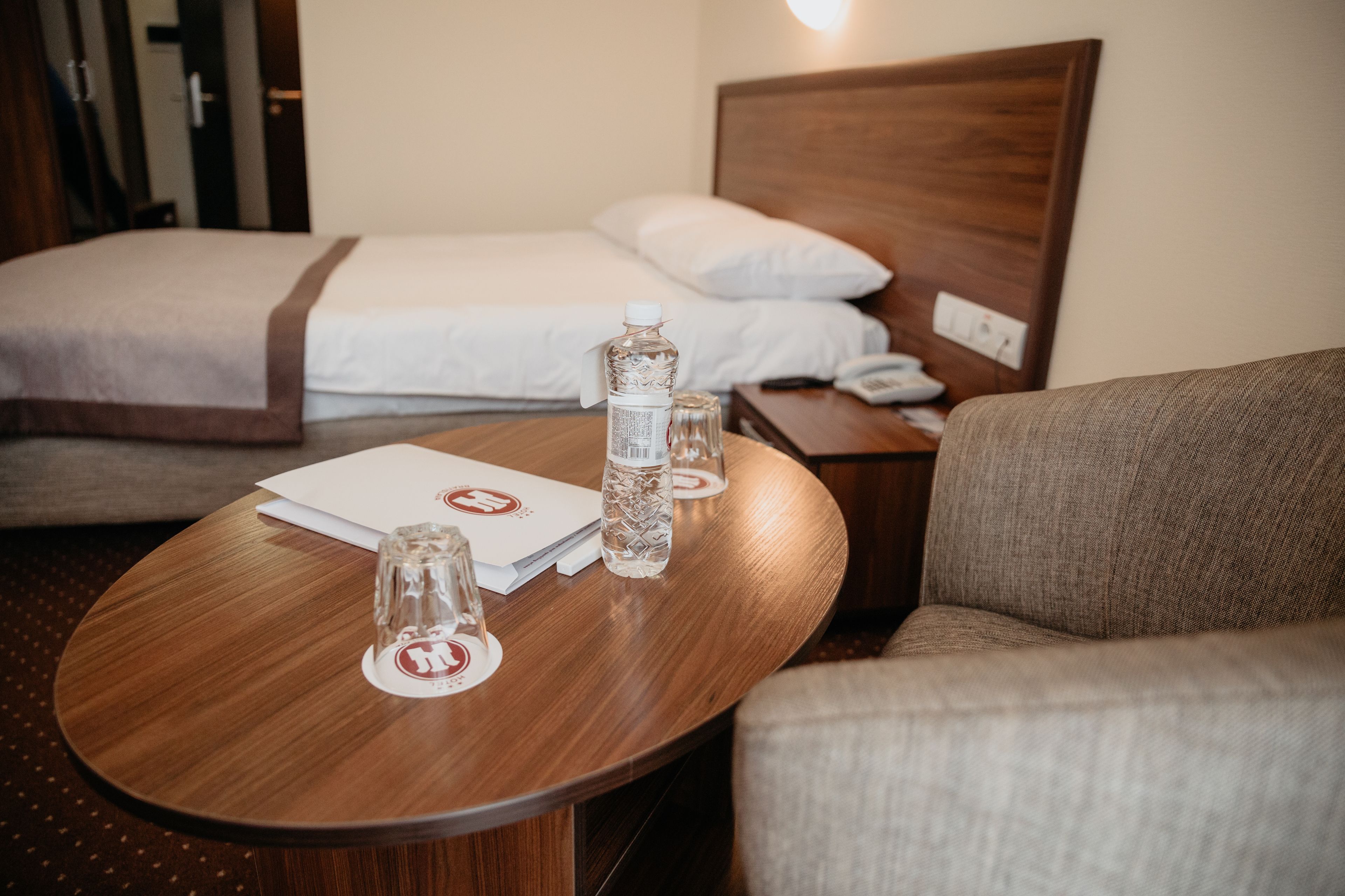 A hotel room with a wooden round table, a bed with white linens, and a grey armchair.