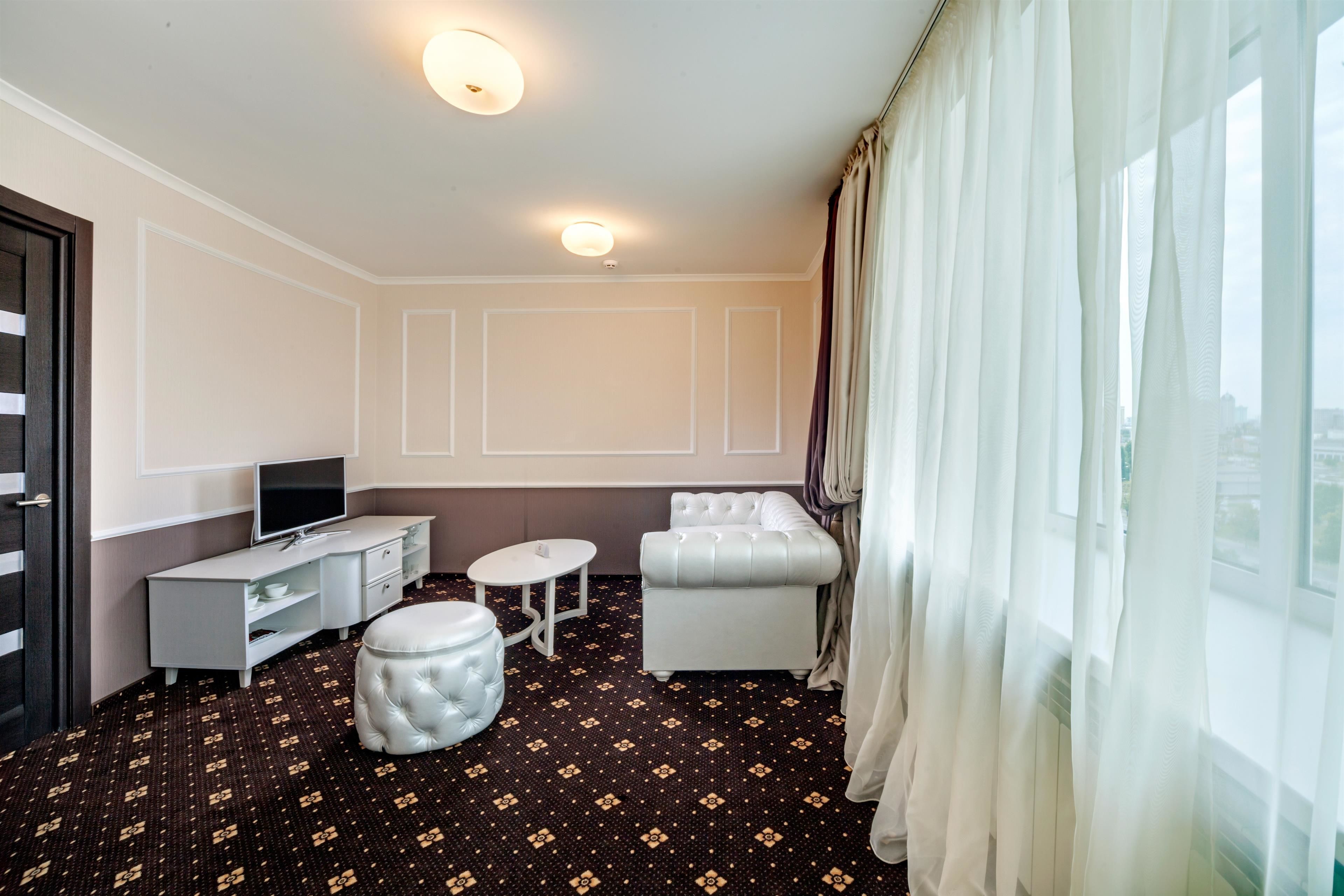 Habitación de hotel con alfombra oscura estampada, un sofá blanco, un sillón blanco, una mesita redonda pequeña, un televisor sobre un mueble blanco y un ventanal con cortinas translúcidas.