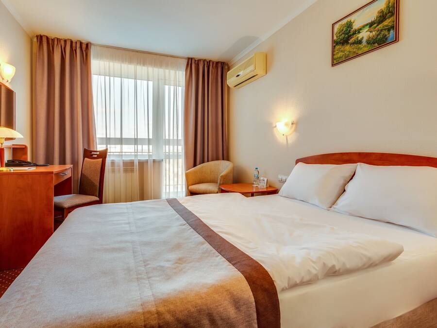 A hotel room with a large bed, a desk with a lamp, a chair, and a small table with a bottle.