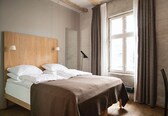 A neatly made bed with white linens, a wooden headboard, and a brown bedspread. There is a window with curtains, a desk with a chair, and a floor lamp.