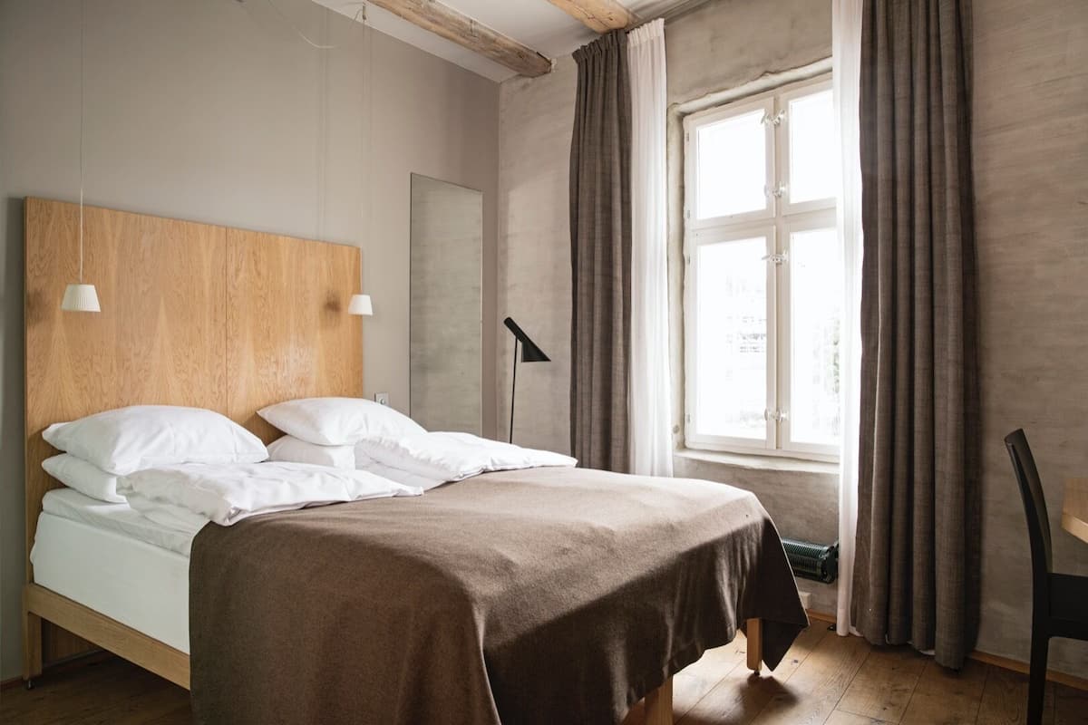 Ein ordentlich bezogenes Bett mit weißen Laken, einem hölzernen Kopfteil und einer braunen Tagesdecke. Es gibt ein Fenster mit Vorhängen, einen Schreibtisch mit Stuhl sowie eine Stehlampe.