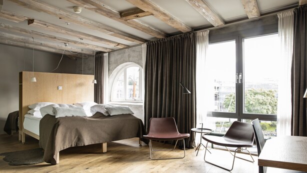 A modern bedroom with a wooden ceiling, a large bed, two chairs, and a view of the city through large windows. - Hotel Brosundet (Ålesund)