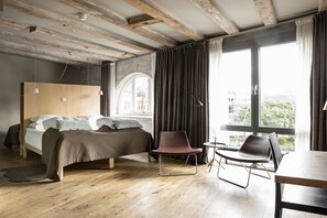 A modern bedroom with a wooden ceiling, a large bed, two chairs, and a view of the city through large windows. - Hotel Brosundet (Ålesund)