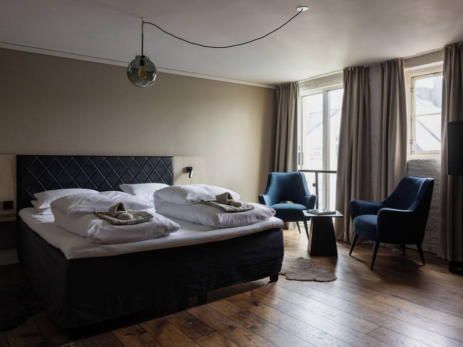 A hotel room with a large bed, two chairs, a small table, and a window with curtains.
