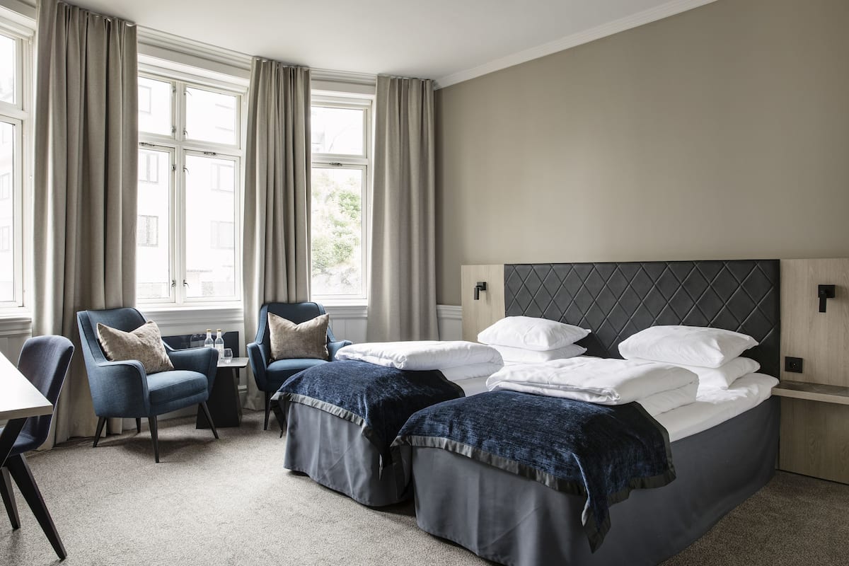 Ein Hotelzimmer mit zwei Betten, einem Schreibtisch, einem Stuhl und großen Fenstern mit Vorhängen.