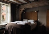 A bedroom with a wooden headboard, a window with curtains, a bedside table with a lamp, and a bed with white linens.