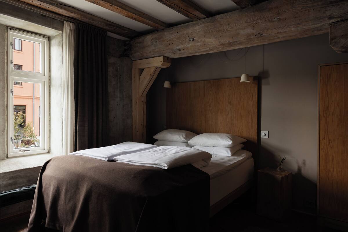 Ein Schlafzimmer mit Holz-Kopfteil, einem fenster mit Vorhängen, einem Nachttisch mit Lampe und einem Bett mit weißen Leinen.