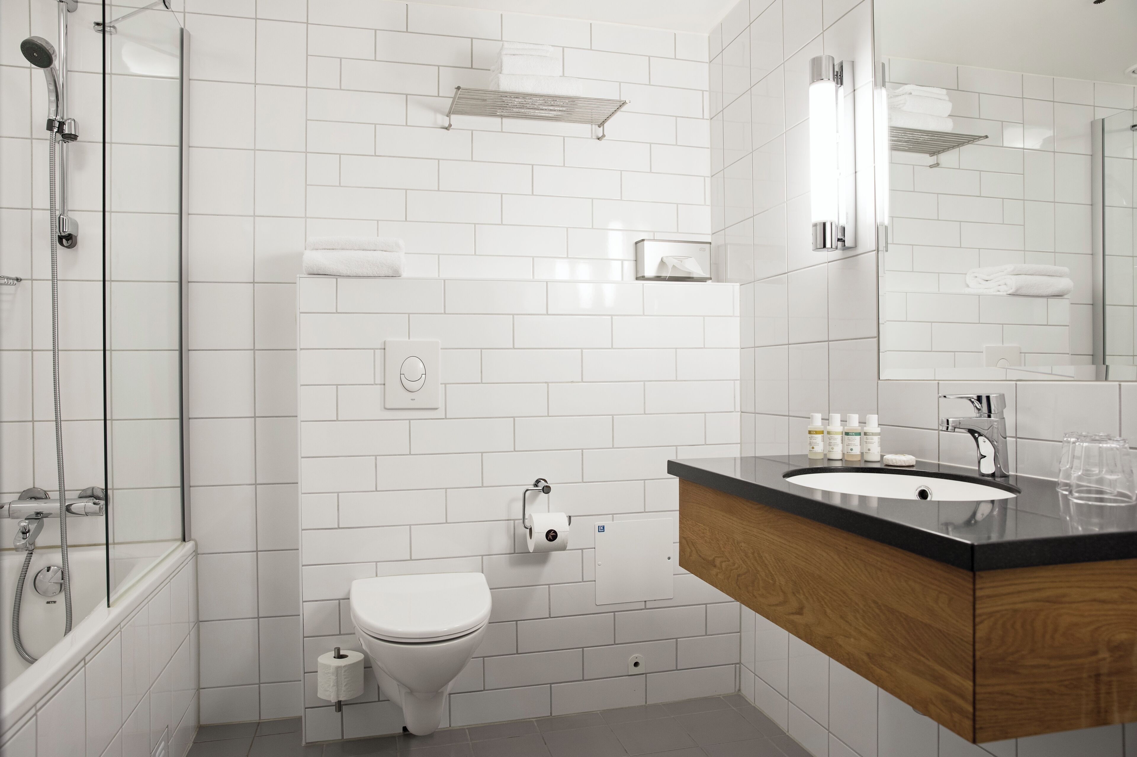 A modern bathroom with white tiles, a wooden vanity, a sink, a toilet, and a shower.