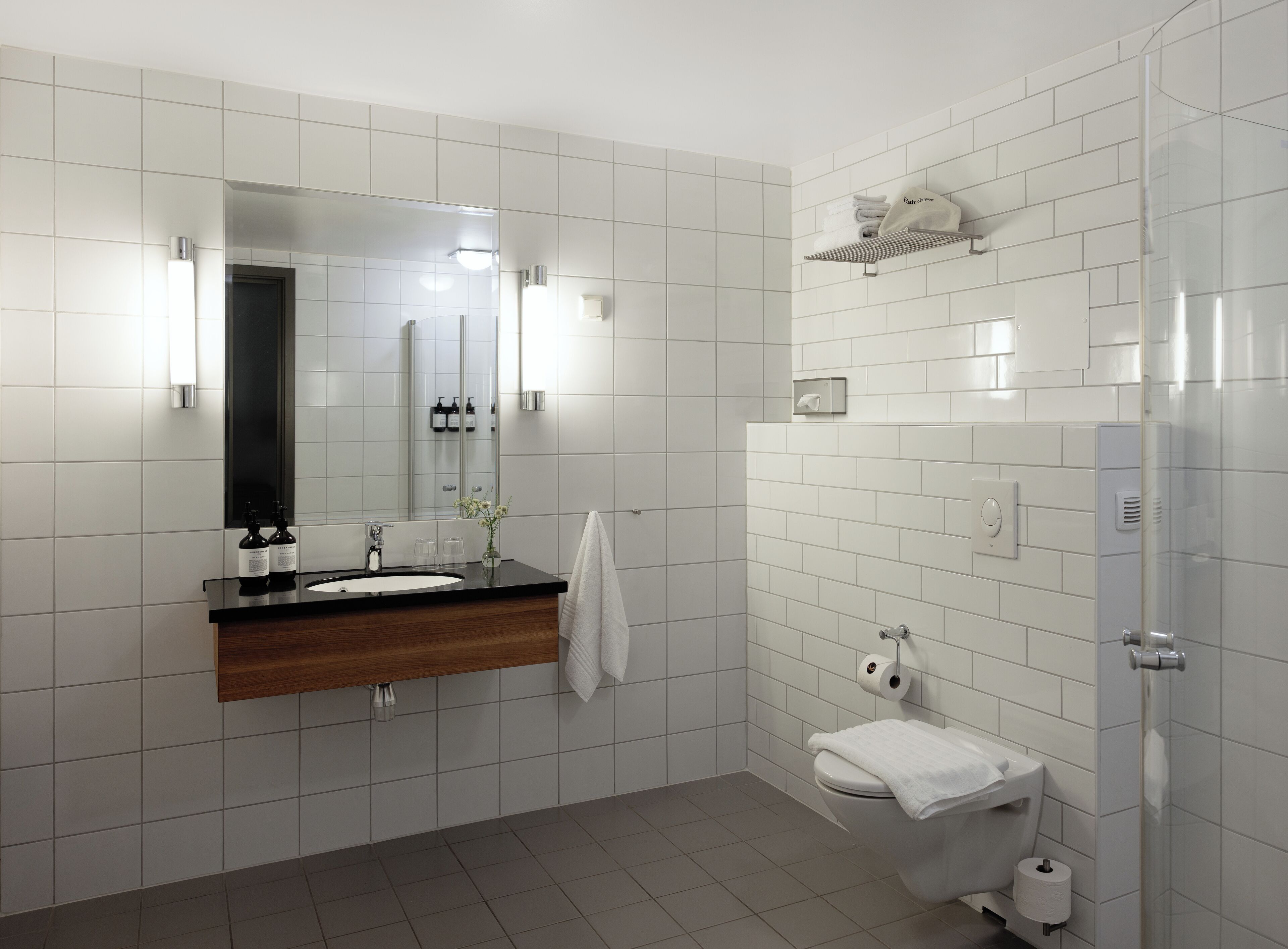 Ein modernes Badezimmer mit weißen Fliesen, einem Holz-Waschtisch, einem Spiegel, einem Handtuchhalter, einer Toilette und einer Duschkabine aus Glas.