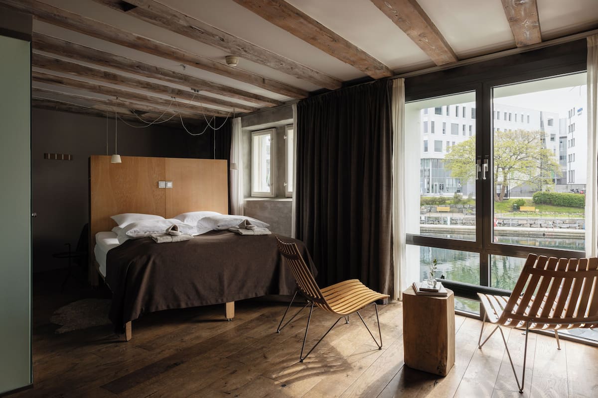 Ein modernes Schlafzimmer mit einem hölzernen Bett, einem großen Fenster und Blick auf ein Gebäude gegenüber dem Wasser.