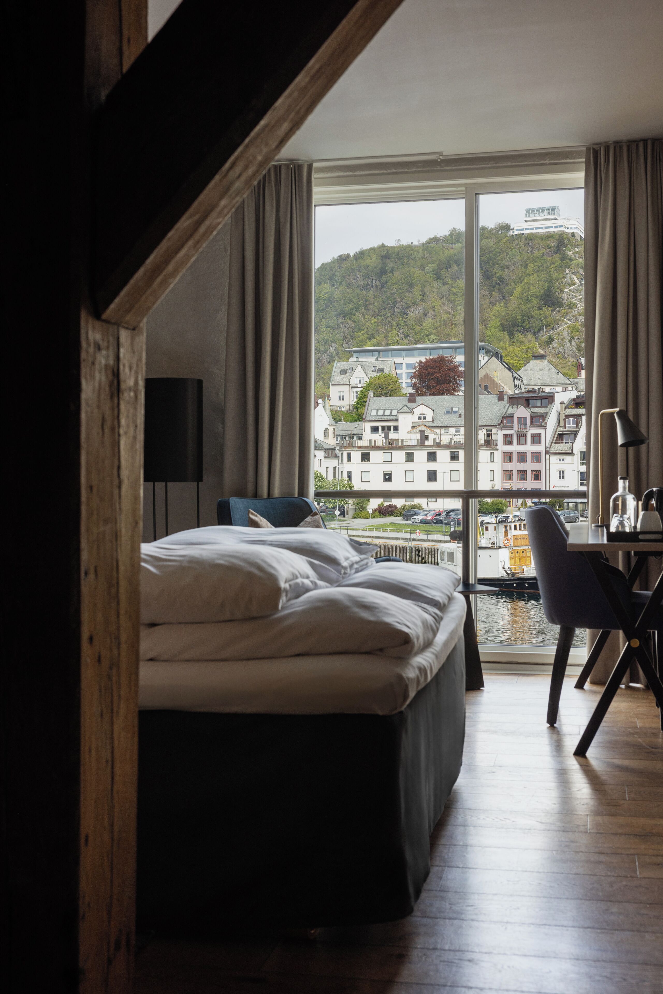 A hotel room with a large bed, a desk, and a view of a town through a large window.