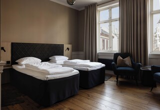 A hotel room with two beds, a chair, a small table, and a view of the city through the window.