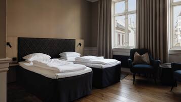 A hotel room with two beds, a chair, a small table, and a view of the city through the window.