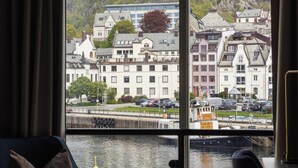 A hotel room with a large window offering a view of a hillside with buildings and a body of water. - Hotel Brosundet (Ålesund)