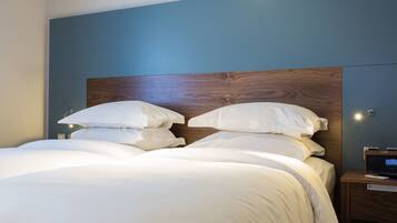 Una cama bien hecha con sábanas blancas y cabecera de madera, una mesita de noche con lámpara y un televisor montado en la pared.