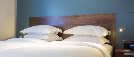 Una cama bien hecha con sábanas blancas y cabecera de madera, una mesita de noche con lámpara y un televisor montado en la pared.