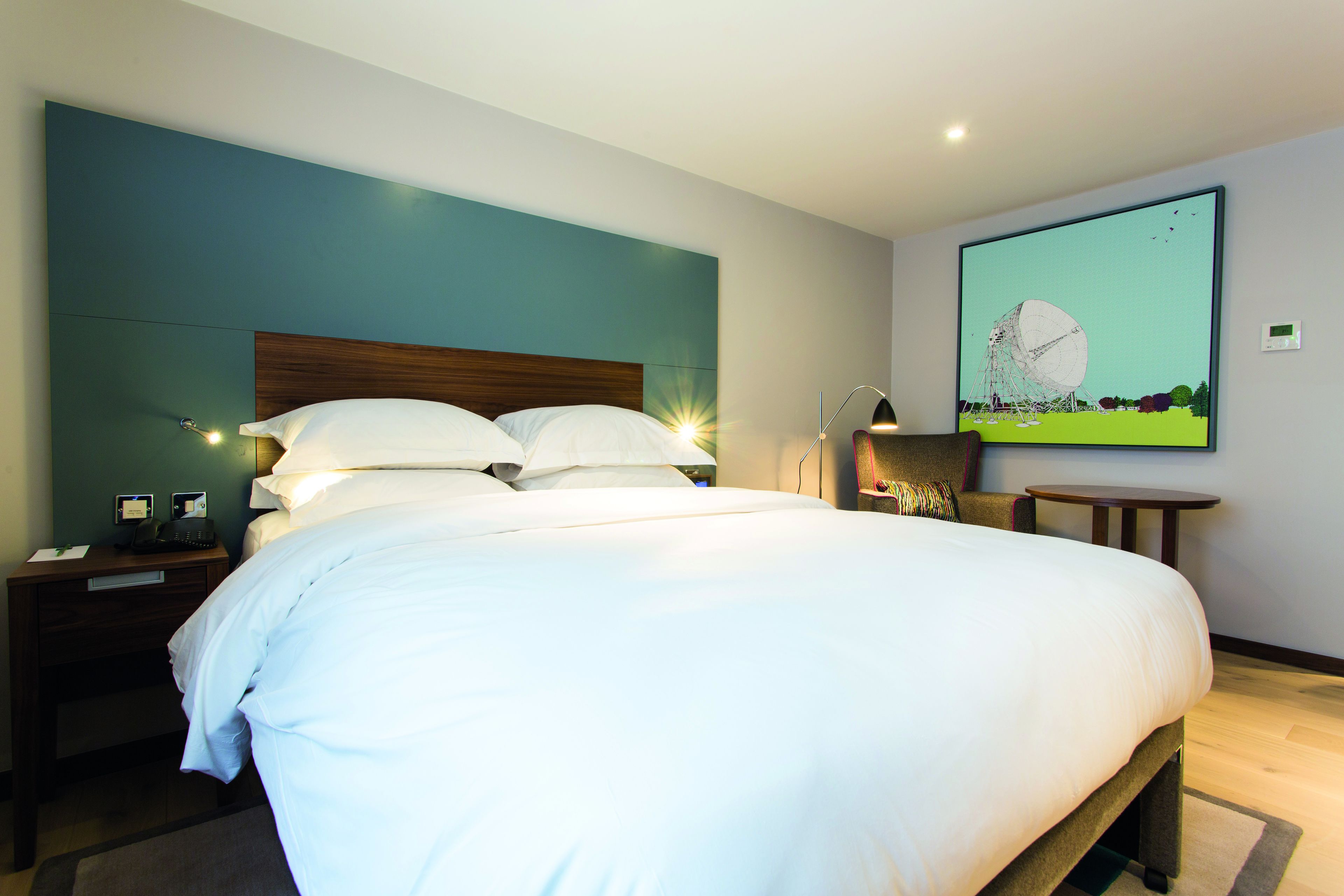 Ein modernes Hotelzimmer mit einem großen Bett, einem Nachttisch mit Telefon und Lampe, einem kleinen Tisch und einem Bild an der Wand.