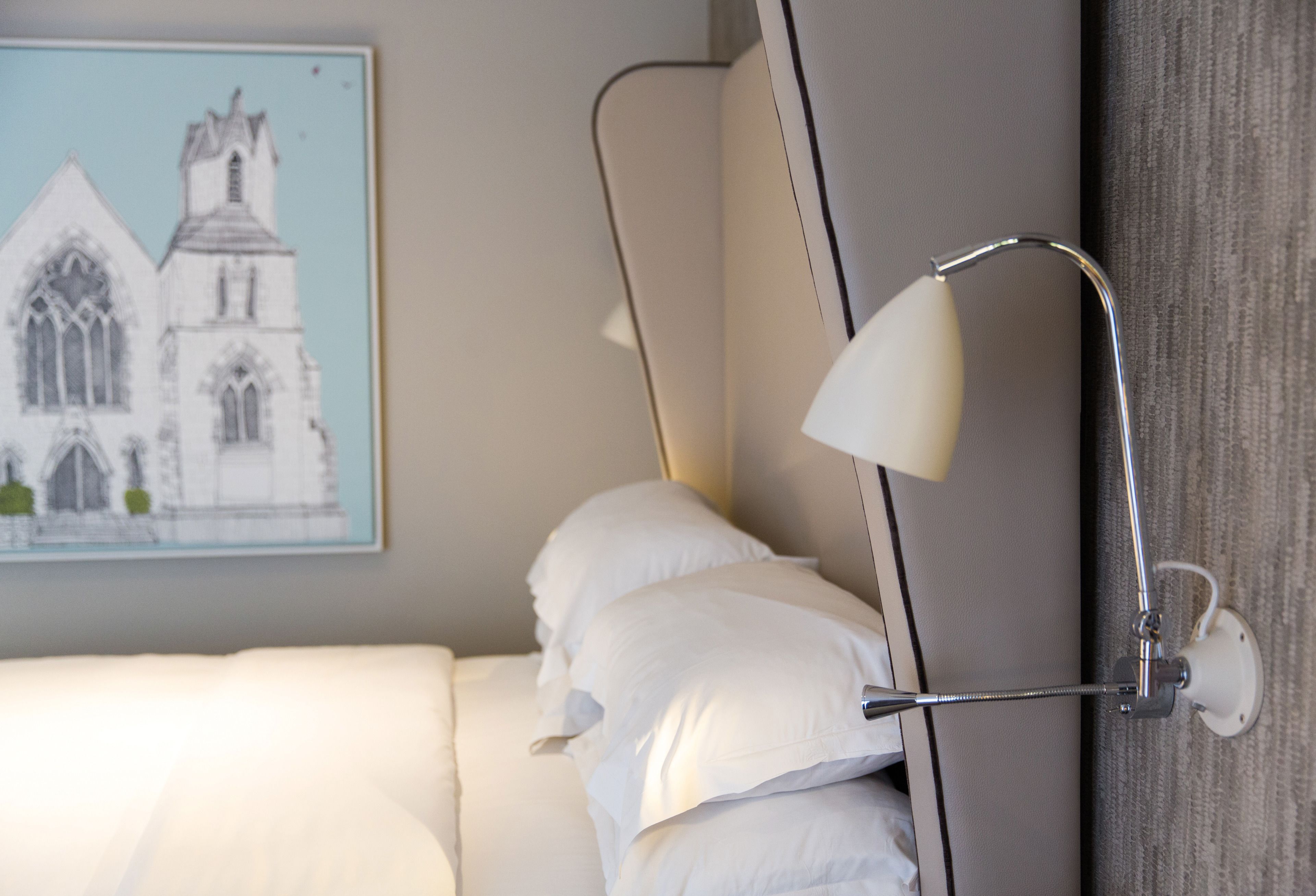 Una cama bien hecha con sábanas blancas, una lámpara fijada a la pared y un cuadro con una iglesia.