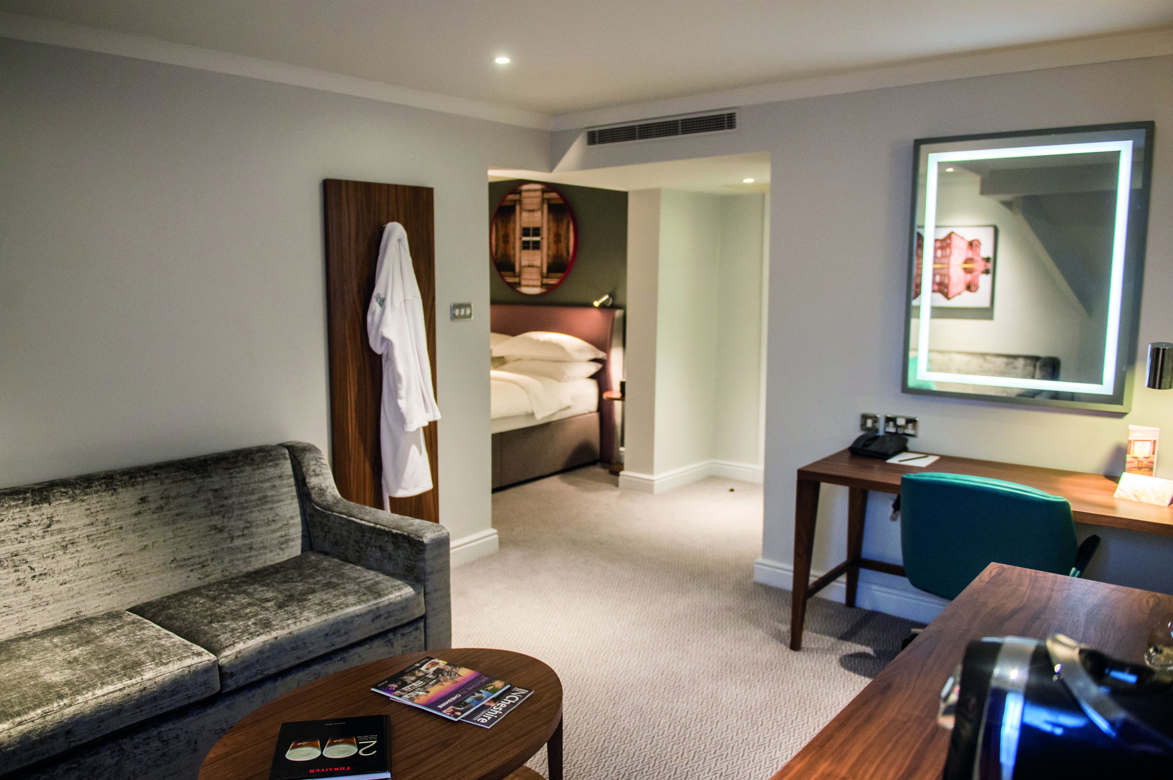 A hotel room with a grey sofa, a small table with a magazine, a desk with a chair, a bed with a headboard, and a large mirror on the wall.
