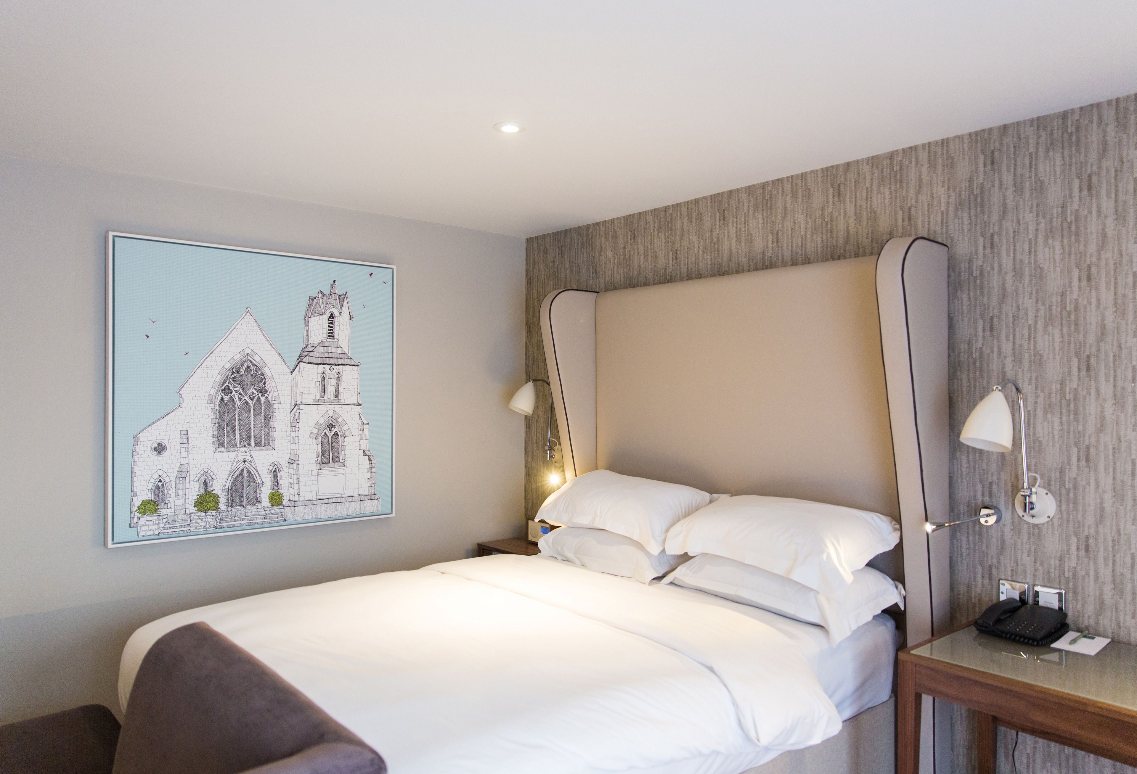 Een hotelkamer met een groot bed, een nachtkastje met een lamp, een lijst met een afbeelding van een kerk en een houten hoofdeinde.