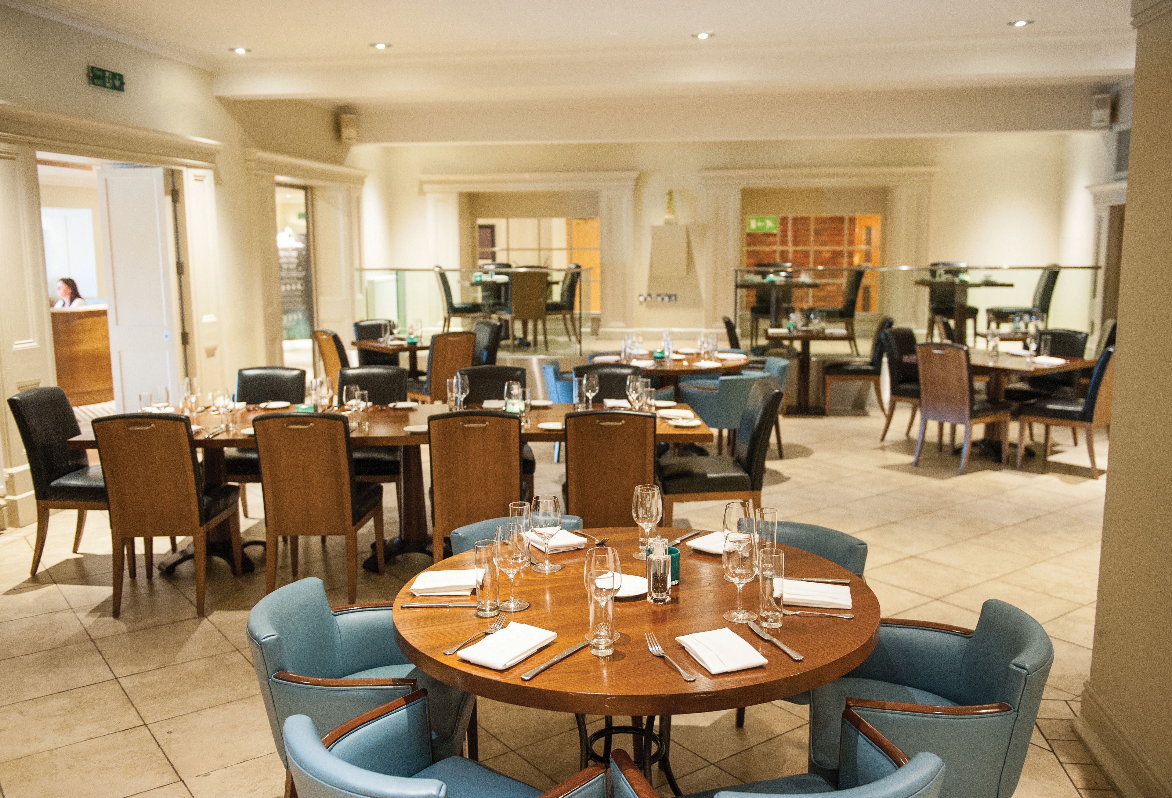 A spacious dining area with multiple round tables and blue chairs, equipped with wine glasses and cutlery.