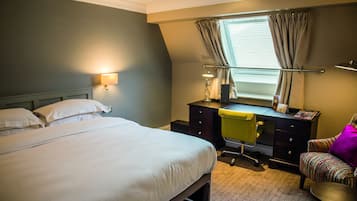 A hotel room with a large bed, a desk with a chair, a desk lamp, a window with curtains, and a small armchair.