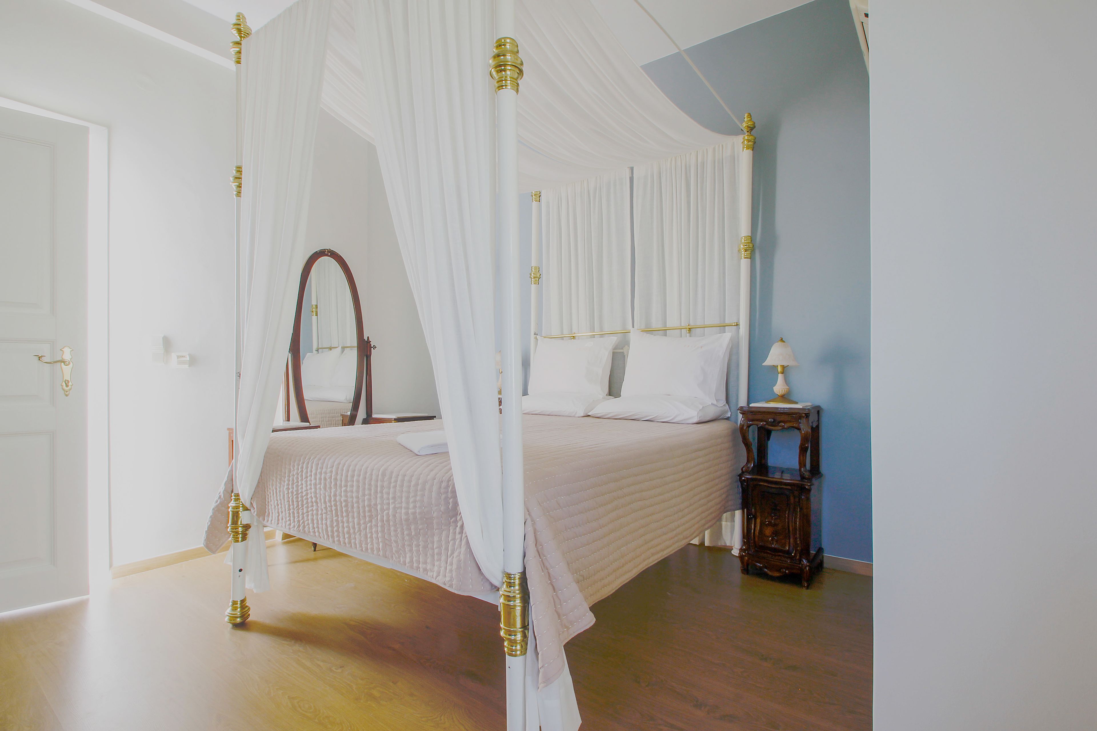A four-poster canopy bed with white linens and gold accents, a bedside table with a lamp, and a large mirror on the wall.
