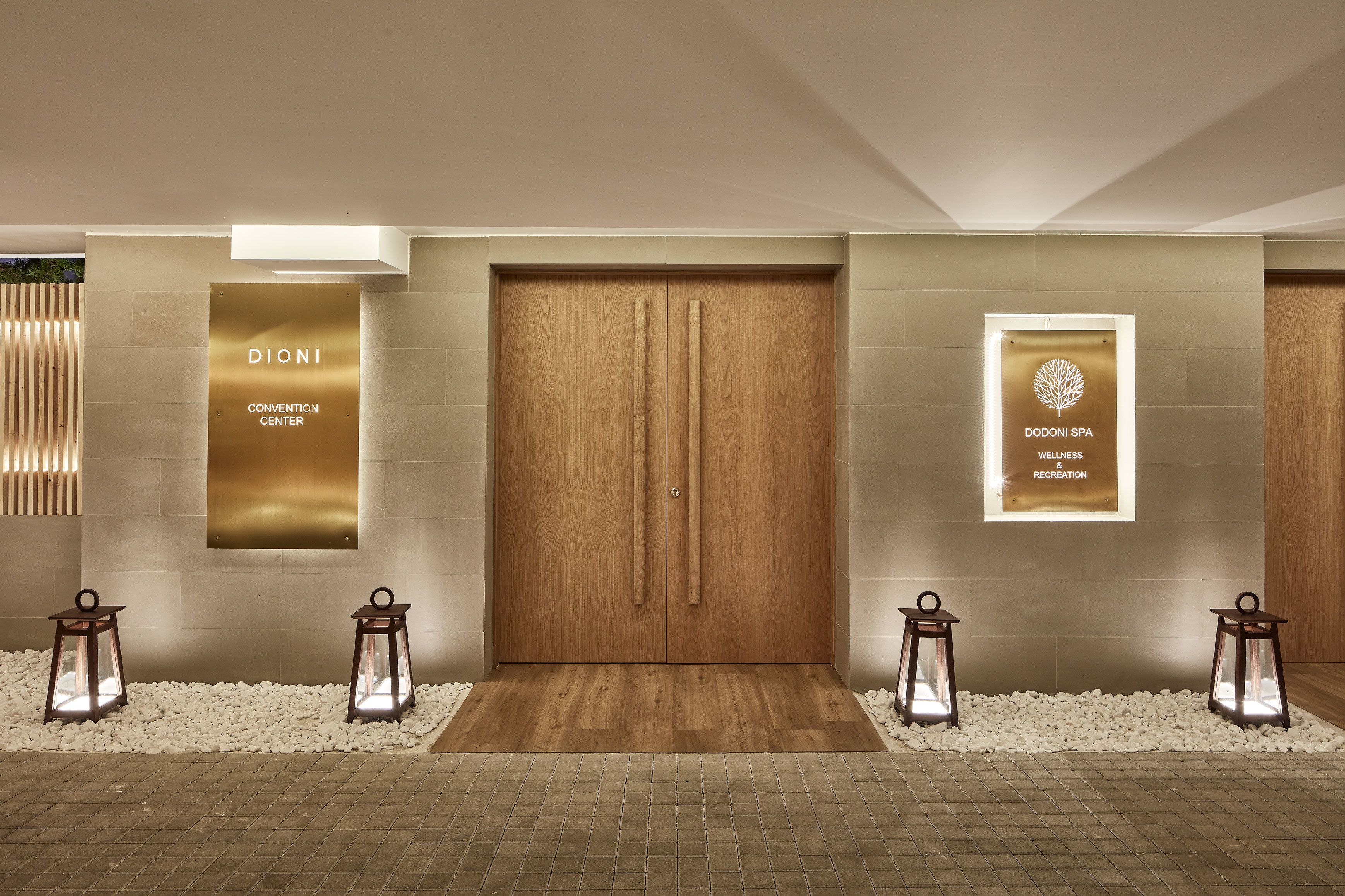 Une entrée moderne avec une porte en bois, deux lanternes et des panneaux indiquant « CENTRE DE CONGRÈS DIONI » et « CENTRE DE BIEN-ÊTRE ET DE LOISIRS DODONI ».