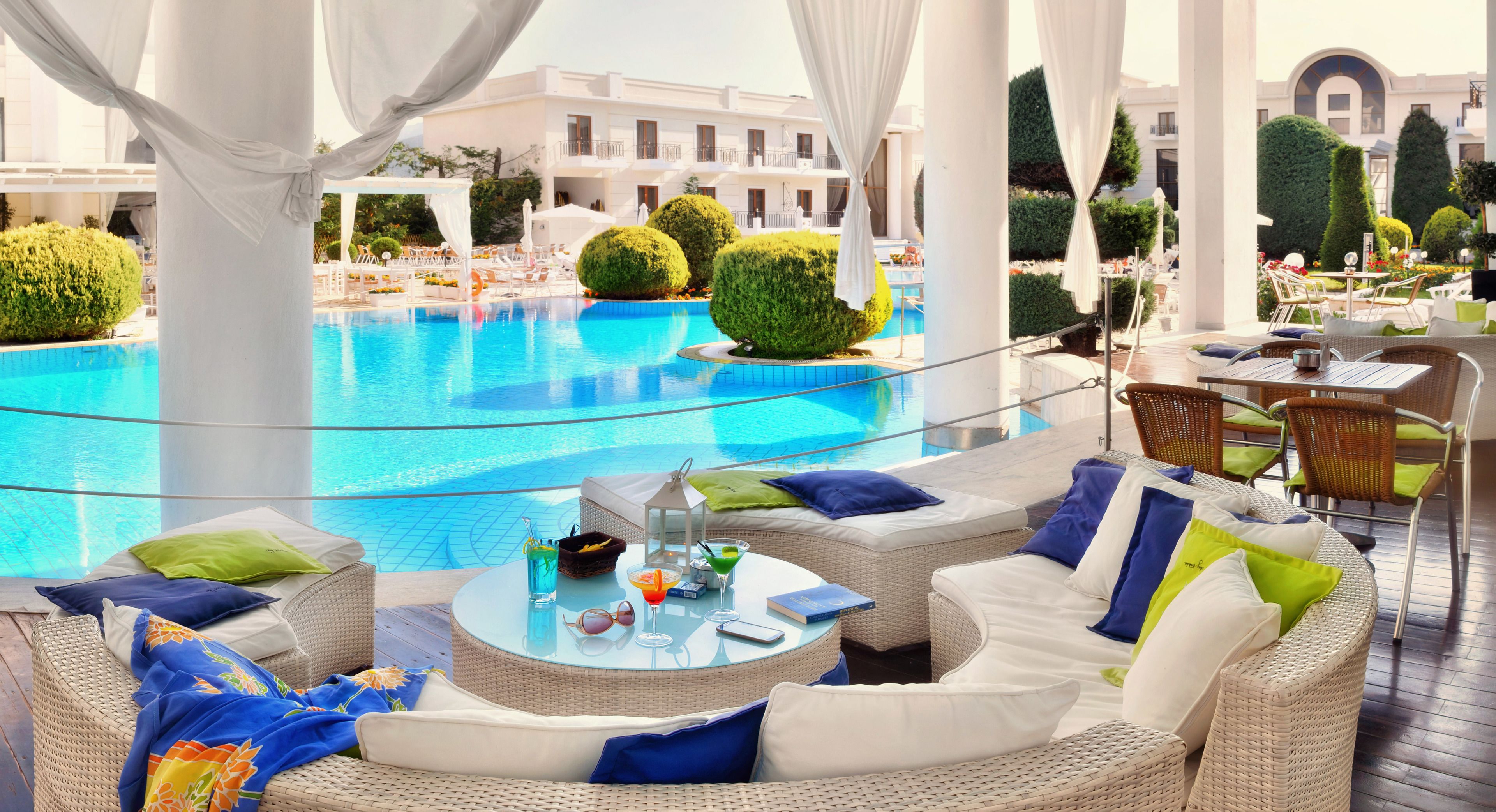 A poolside lounge with wicker furniture, a glass table, and a view of the pool and hotel buildings.