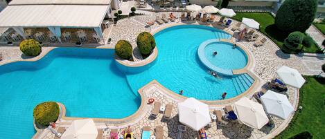 Una piscina grande con varias zonas de sol y sombrillas, rodeada por una terraza de piedra y follaje.