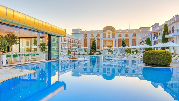 A hotel pool area with a clear pool, sun loungers, umbrellas, and a building with multiple balconies. - Epirus Palace Congress & Spa (Ioannina)