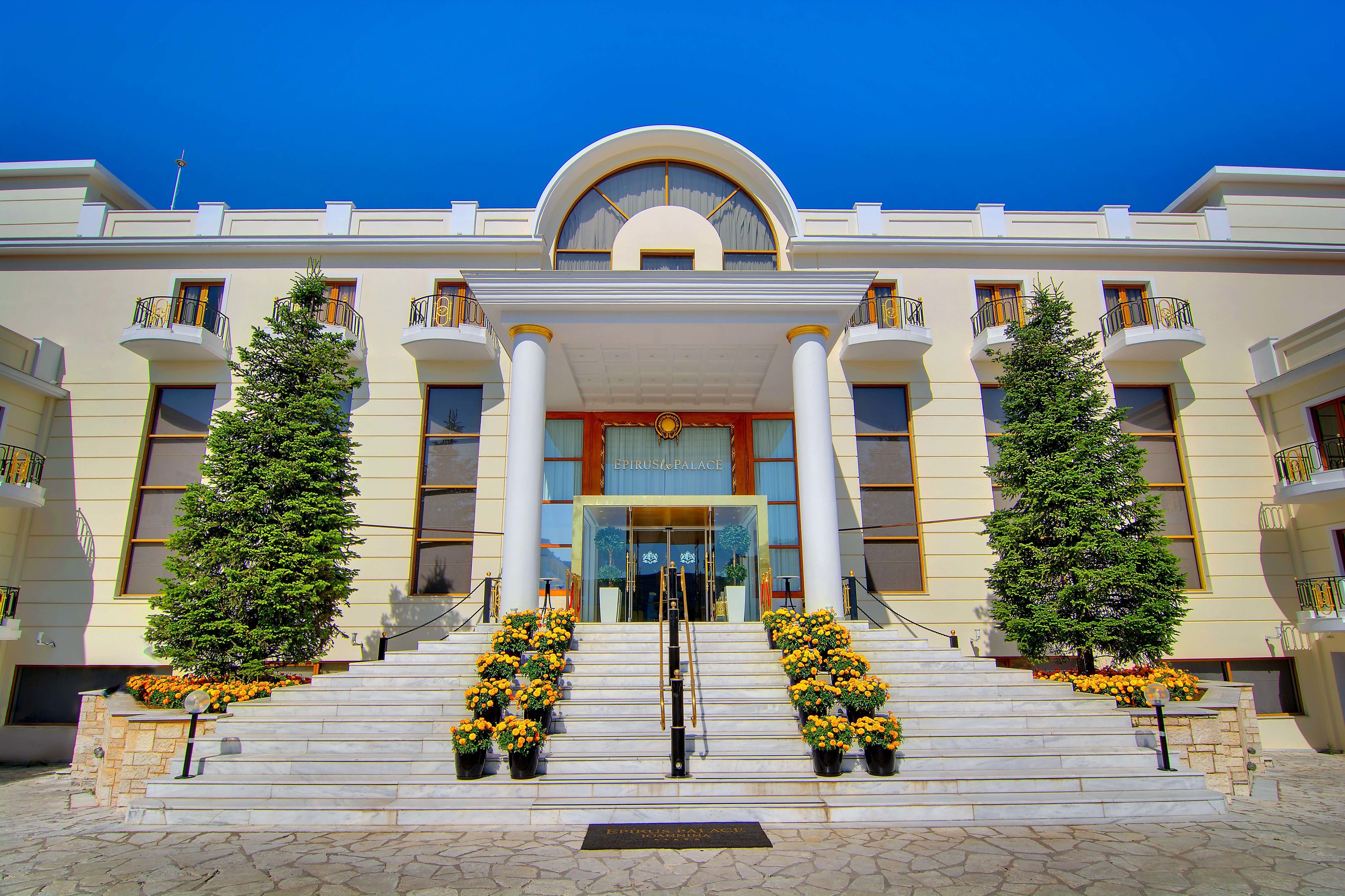 A grand hotel entrance with a symmetrical facade, large columns, and a central entrance flanked by two trees and flower arrangements.
