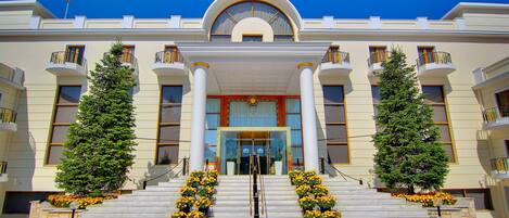 A grand hotel entrance with a symmetrical facade, large columns, and a central entrance flanked by two trees and flower arrangements.